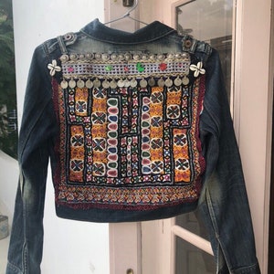 Puede incluir: Chaqueta vaquera con intrincados bordados y adornos. La parte trasera de la chaqueta presenta un colorido patr&oacute;n geom&eacute;trico en rojo, amarillo y negro. Monedas plateadas y elementos decorativos adornan la parte superior de la espalda. La chaqueta est&aacute; colgada de una percha met&aacute;lica.