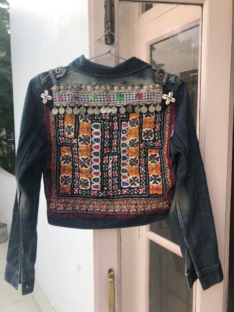 Puede incluir: Chaqueta vaquera con intrincados bordados y adornos. La parte trasera de la chaqueta presenta un colorido patr&oacute;n geom&eacute;trico en rojo, amarillo y negro. Monedas plateadas y elementos decorativos adornan la parte superior de la espalda. La chaqueta est&aacute; colgada de una percha met&aacute;lica.