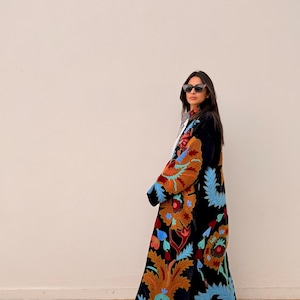 May include: A long, black velvet coat adorned with colourful floral embroidery in orange, blue, and red. The coat has a flowing design and detailed patterns, creating a bohemian style. This statement coat is ideal for adding an artistic touch to any outfit.