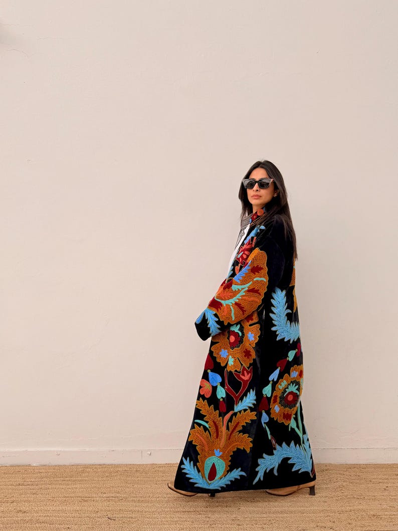 May include: A long, black velvet coat adorned with colourful floral embroidery in orange, blue, and red. The coat has a flowing design and detailed patterns, creating a bohemian style. This statement coat is ideal for adding an artistic touch to any outfit.