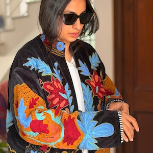 May include: A black velvet jacket with colourful floral embroidery in orange, red, and blue. The jacket has a mandarin collar and a white lining. The sleeves have a black and white striped cuff.