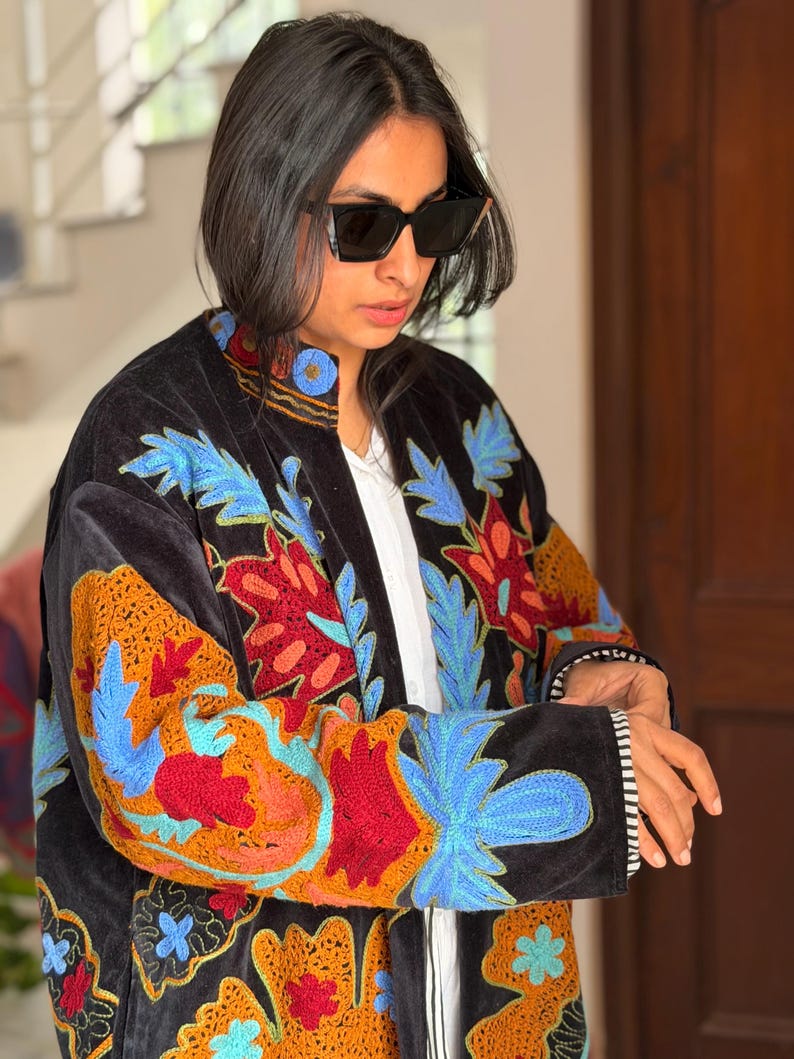 May include: A black velvet jacket with colourful floral embroidery in orange, red, and blue. The jacket has a mandarin collar and a white lining. The sleeves have a black and white striped cuff.