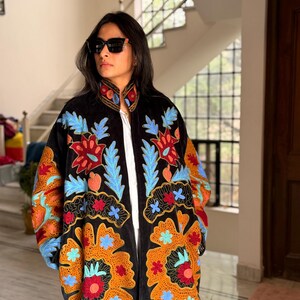 May include: Black embroidered coat with a vibrant floral design in shades of orange, red, blue, and green. The coat features a high collar and long sleeves. The embroidery is detailed and covers the front and sleeves.