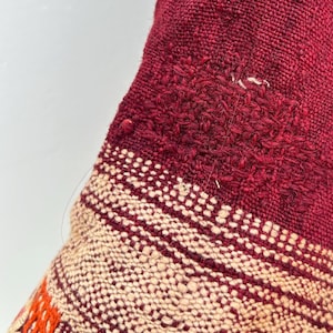 May include: Close-up of a red and cream woven fabric with colorful embroidery. The fabric has a textured, hand-woven look and features a geometric pattern.
