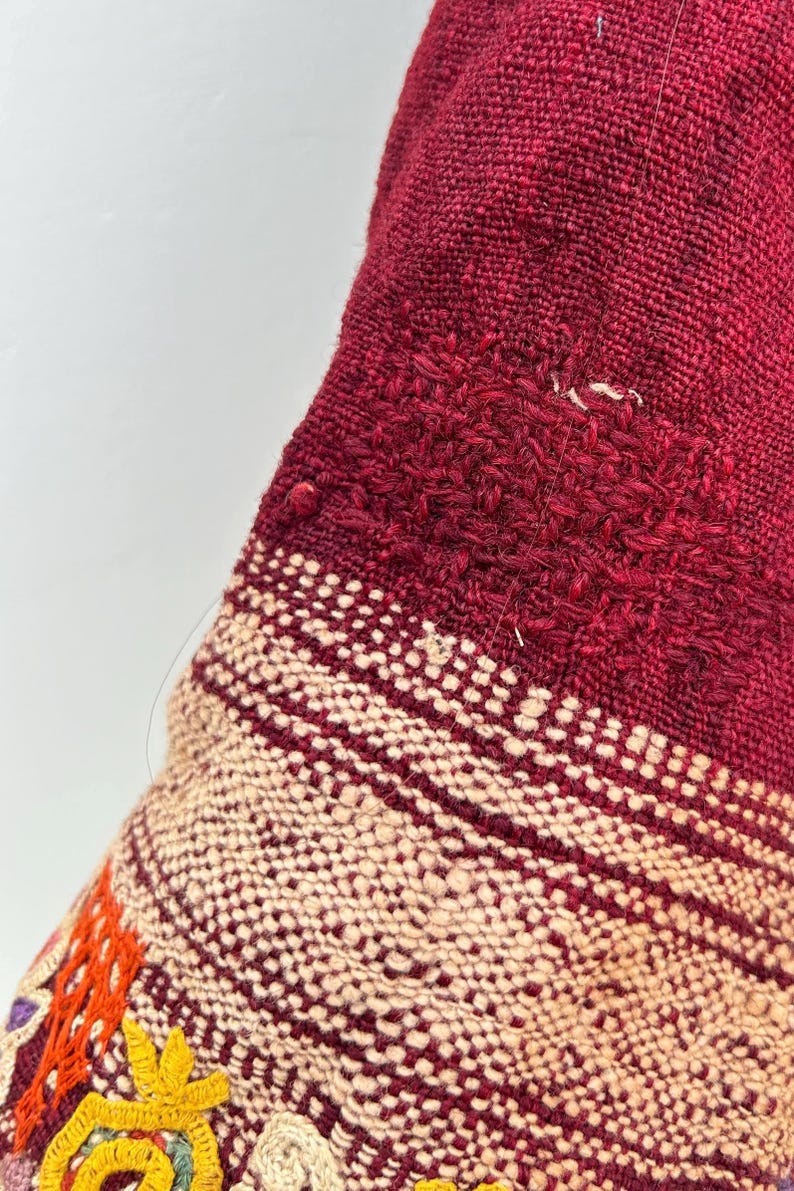 May include: Close-up of a red and cream woven fabric with colorful embroidery. The fabric has a textured, hand-woven look and features a geometric pattern.
