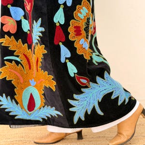 May include: Black velvet skirt with vibrant floral embroidery in orange, red, blue, and green. The skirt showcases an intricate design, a white hemline, and is worn with brown heeled boots.