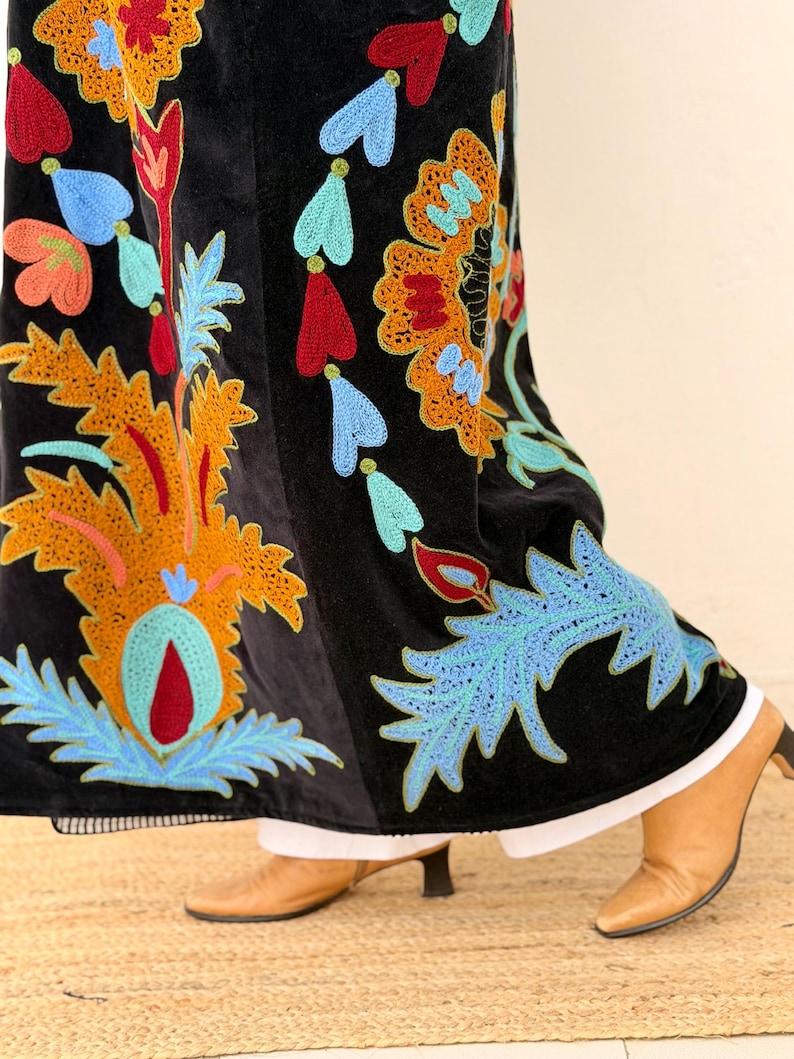 May include: Black velvet skirt with vibrant floral embroidery in orange, red, blue, and green. The skirt showcases an intricate design, a white hemline, and is worn with brown heeled boots.