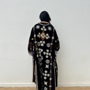 May include: A long, black embroidered coat with white floral and geometric patterns. The coat features brown and white trim, and a diamond-shaped design on the upper back. The sleeves have striped cuffs. The coat is worn by a person.