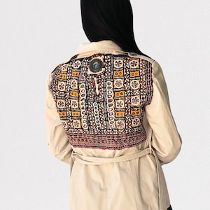 Può includere: Una giacca utility beige con un pannello posteriore ricamato colorato. La giacca ha una cintura e maniche lunghe.