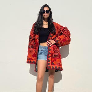 Puede incluir: Una mujer con una chaqueta larga de color rojo y negro con un diseño geométrico. La chaqueta tiene un cuello chal y mangas largas. También lleva pantalones cortos de mezclilla y botas marrones.