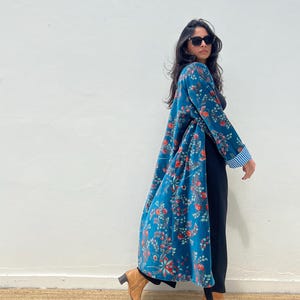 May include: A long, flowing teal floral print robe with red and orange flowers. The robe has a blue and white striped cuff. The person is wearing black pants and brown boots.