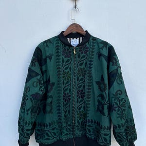 Puede incluir: Una chaqueta bomber verde con un estampado floral negro y cierre de cremallera. La chaqueta tiene un cuello, puños y dobladillo de canalé negros. La etiqueta de la chaqueta dice "AL MRO".