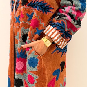Puede incluir: Un abrigo largo de color &oacute;xido con intrincados bordados florales en tonos rosa, azul, negro y gris. El abrigo tiene un bolsillo y pu&ntilde;os a rayas. Se ve un reloj de oro en la mu&ntilde;eca.