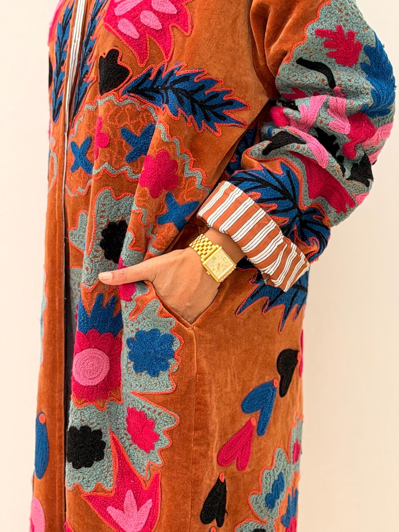 Puede incluir: Un abrigo largo de color &oacute;xido con intrincados bordados florales en tonos rosa, azul, negro y gris. El abrigo tiene un bolsillo y pu&ntilde;os a rayas. Se ve un reloj de oro en la mu&ntilde;eca.