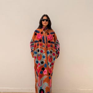 Puede incluir: Un abrigo largo de color &oacute;xido con intrincados bordados florales en tonos rosa, azul y verde. El abrigo presenta un cuello, mangas largas y un dise&ntilde;o de cuerpo entero. El bordado crea un contraste llamativo.