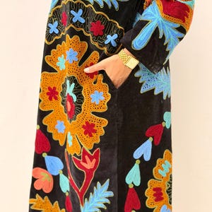 May include: Black velvet coat with colourful floral embroidery in orange, blue, red, and green. The coat has a long, flowing design with intricate patterns and a pocket. The sleeves and front panels are adorned with detailed stitching.