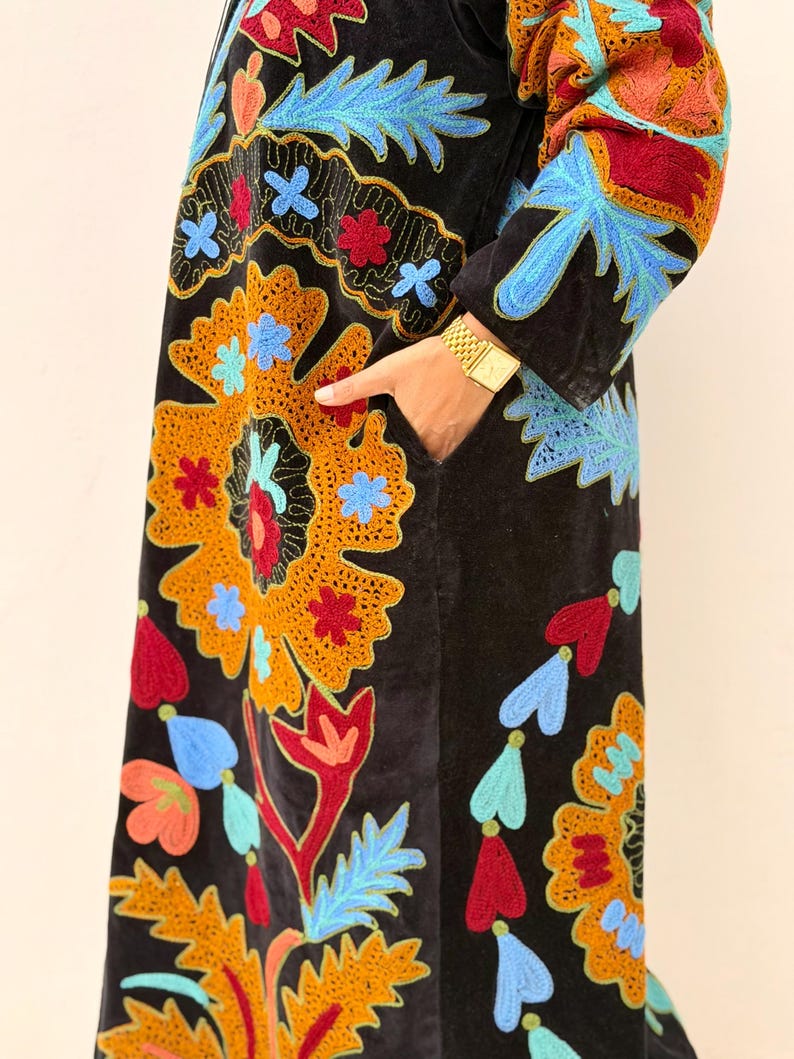 May include: Black velvet coat with colourful floral embroidery in orange, blue, red, and green. The coat has a long, flowing design with intricate patterns and a pocket. The sleeves and front panels are adorned with detailed stitching.