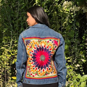 May include: A blue denim jacket with a colorful embroidered patch on the back. The patch features a large, multi-colored flower design with intricate details.