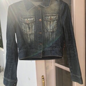 Puede incluir: Chaqueta vaquera azul oscuro con cuello cl&aacute;sico, cierre de botones y dos bolsillos en el pecho. La chaqueta tiene un aspecto ligeramente descolorido y est&aacute; colgada de una percha met&aacute;lica.