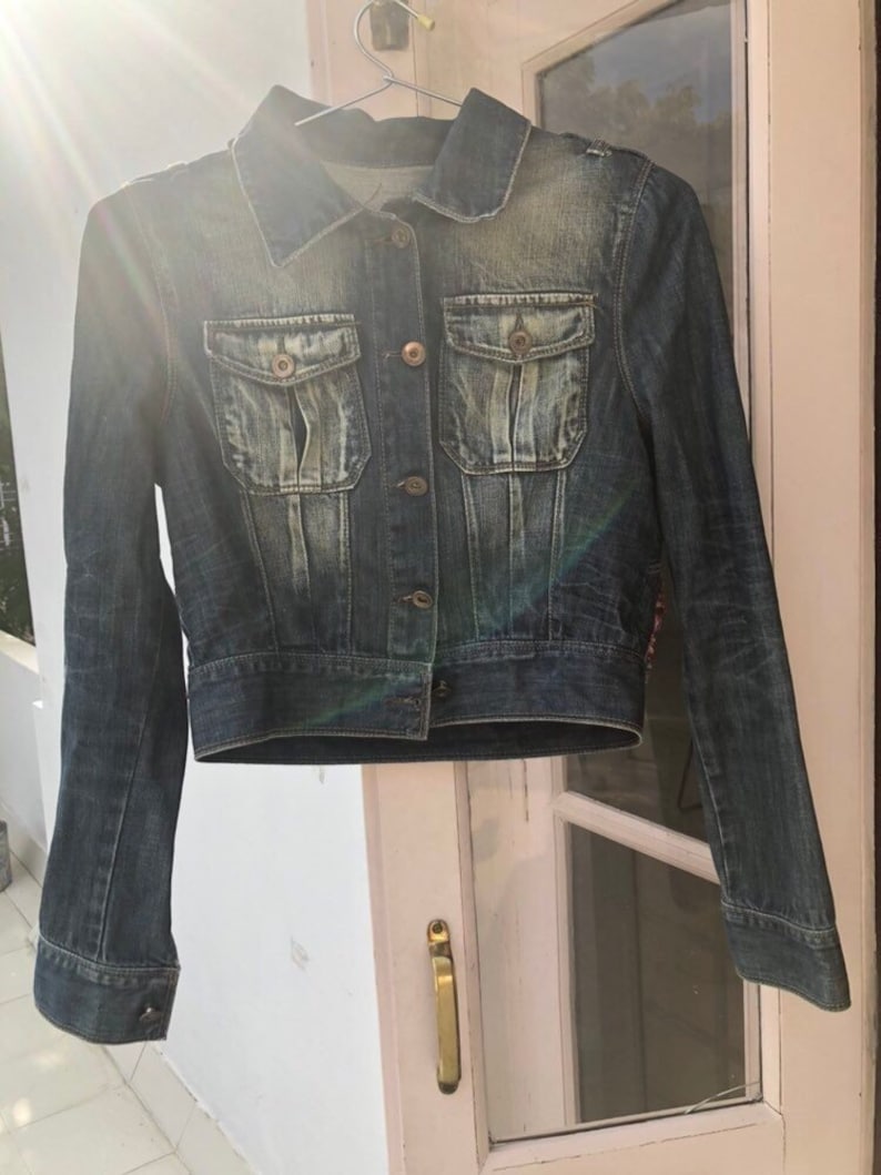 Puede incluir: Chaqueta vaquera azul oscuro con cuello cl&aacute;sico, cierre de botones y dos bolsillos en el pecho. La chaqueta tiene un aspecto ligeramente descolorido y est&aacute; colgada de una percha met&aacute;lica.