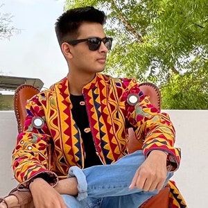 Puede incluir: Una persona lleva una chaqueta estampada en amarillo, rojo y azul con adornos de espejo. La chaqueta tiene un cuello alto y cierre de botón. La persona lleva pantalones vaqueros azules y gafas de sol.