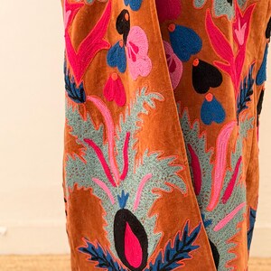 Puede incluir: Prenda de terciopelo color &oacute;xido con vibrantes bordados florales. El bordado presenta dise&ntilde;os de flores y hojas rosas, azules y negras. La textura y el color de la prenda sugieren un estilo bohemio lujoso, adecuado para combinar.