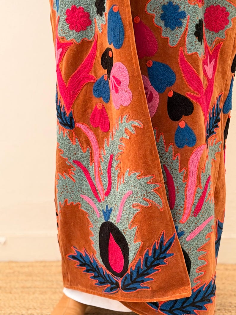 Puede incluir: Prenda de terciopelo color &oacute;xido con vibrantes bordados florales. El bordado presenta dise&ntilde;os de flores y hojas rosas, azules y negras. La textura y el color de la prenda sugieren un estilo bohemio lujoso, adecuado para combinar.