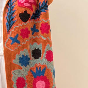 Puede incluir: Un abrigo naranja quemado con bordados florales y de hojas coloridas. El bordado presenta detalles en rosa, azul y negro, creando un patr&oacute;n vibrante y detallado. La textura del abrigo parece suave y afelpada.