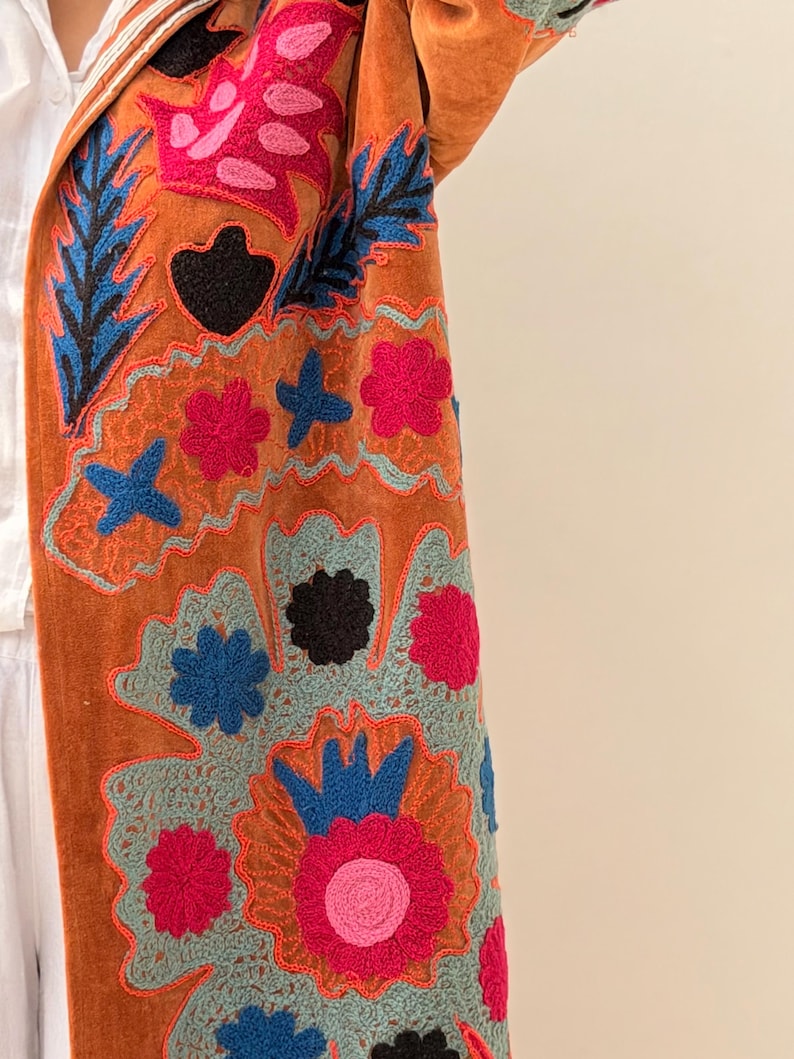 Puede incluir: Un abrigo naranja quemado con bordados florales y de hojas coloridas. El bordado presenta detalles en rosa, azul y negro, creando un patr&oacute;n vibrante y detallado. La textura del abrigo parece suave y afelpada.