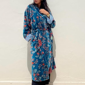 May include: A teal velvet robe with a floral pattern in red and pink. The robe has a matching belt and striped cuffs. The person is wearing black sunglasses and tan boots. The robe is a stylish piece of clothing.