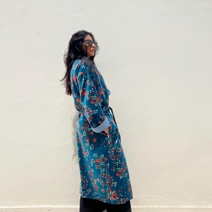 May include: A long, teal floral robe with red and orange flowers. The robe has a belt, pockets, and blue and white striped cuffs. The person is wearing black pants and tan ankle boots.