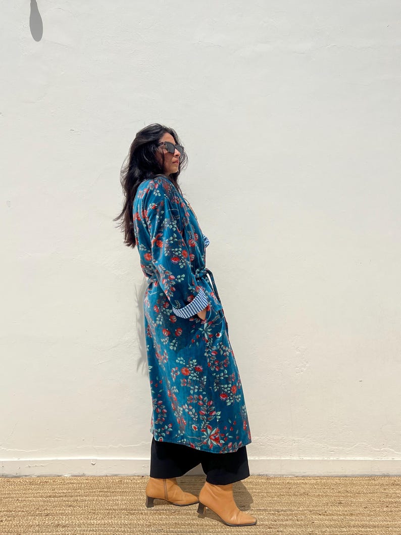 May include: A long, teal floral robe with red and orange flowers. The robe has a belt, pockets, and blue and white striped cuffs. The person is wearing black pants and tan ankle boots.