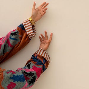 Puede incluir: Chaqueta de terciopelo marr&oacute;n con bordados florales coloridos en rosa, azul y negro. Los pu&ntilde;os presentan un estampado a rayas marr&oacute;n y blanco. Se ve un reloj dorado en la mu&ntilde;eca.
