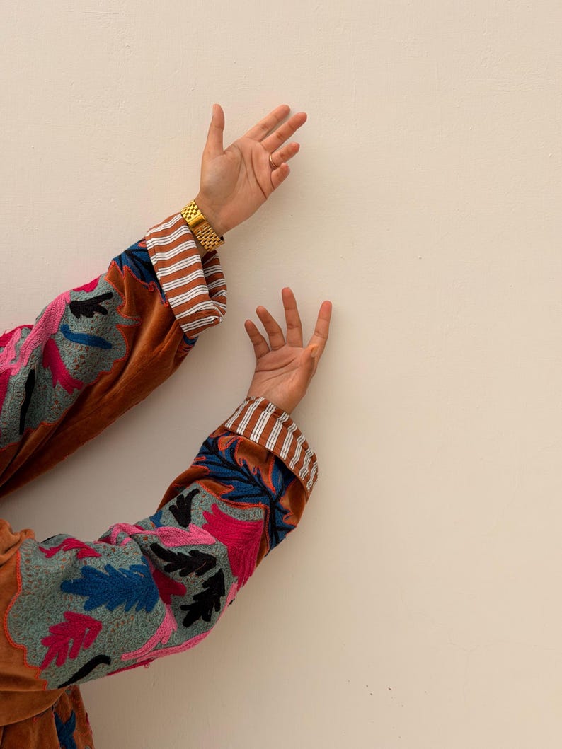 Puede incluir: Chaqueta de terciopelo marr&oacute;n con bordados florales coloridos en rosa, azul y negro. Los pu&ntilde;os presentan un estampado a rayas marr&oacute;n y blanco. Se ve un reloj dorado en la mu&ntilde;eca.