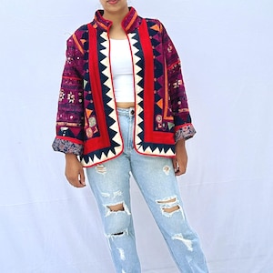 May include: A woman wearing a red, black, and white patterned jacket with a white top and light blue jeans. She is wearing white sneakers.