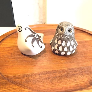 Puede incluir: Dos figuras de pájaros de cerámica. Una es blanca con un diseño floral negro, la otra es un pequeño búho con un patrón moteado en tonos marrones y blancos. Ambas están sobre una bandeja de madera.