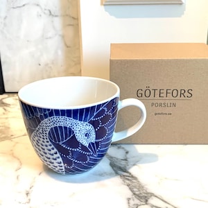 May include: A blue and white ceramic mug featuring a stylised bird design. The mug is made by Götefors Porslin, a Swedish pottery company.