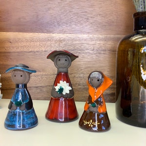 Puede incluir: Tres figuritas de cerámica de personas que llevan sombreros y vestidos. La figurita de la izquierda lleva un vestido y un sombrero azules. La figurita del centro lleva un vestido y un sombrero rojos. La figurita de la derecha lleva un vestido y un sombrero naranjas.