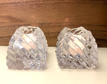 May include: Two clear glass candle holders with a textured, geometric design. The holders are empty and have a square base.