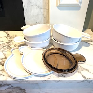 Op de afbeelding: Een set witte keramische soepkommen met handvatten, in paren gestapeld. De kommen worden vergezeld door witte deksels en een bruin glazen deksel. De items zijn gerangschikt op een marmeren oppervlak.