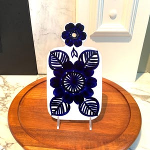 Puede incluir: Tabla de cortar de cerámica blanca con un diseño floral azul. La tabla presenta una gran flor central y hojas circundantes, todo en un tono azul oscuro. La tabla se exhibe sobre una bandeja de madera.