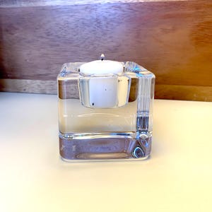 May include: A clear glass candle holder with a white candle inside. The holder has a square shape and a textured surface.