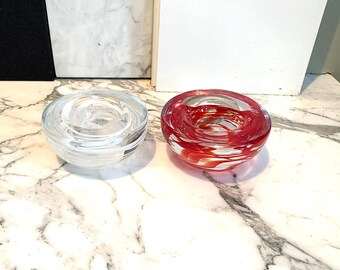May include: Two glass candle holders. One is clear, the other is red with white swirls. Both are round with a spiral design. The candle holders are on a marble surface.