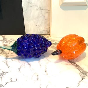 May include: Two glass fruit sculptures. One is a cluster of deep blue grapes with a green stem. The other is an orange pumpkin with yellow spots and a black stem. Both are on a marble surface.