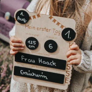 May include: A wooden sign with a chalkboard background and a black and white banner design. The sign says "Mein erster Tag" (My first day) and includes the date "1. Aug." (August 1st), the child's height "125 cm", age "6", teacher's name "Frau Haack", and profession "Erzieherin" (Educator).