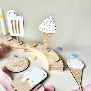 May include: Wooden ice cream cone, popsicle, and ice cream sandwich ornaments with white frosting and sprinkles. The ornaments are on a wooden base with the text "frilly designs" on a round wooden tag.
