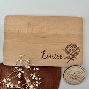 Peut inclure: Planche &agrave; d&eacute;couper rectangulaire en bois clair, grav&eacute;e du pr&eacute;nom "Louise" et d'un motif floral. Le bois est de couleur claire et les coins sont arrondis. Un petit dessous de verre rond en bois avec "Frilly Designs".