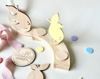 May include: Wooden lemon-shaped candle holder with a yellow lemon cutout and a pink ribbon. The candle holder is on a wooden base with the text "bailey designs" on it.