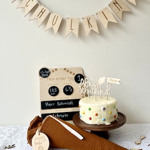 May include: A white cake with colorful sprinkles and a wooden 'Schulkind' cake topper. The cake is on a wooden cake stand. There is a chalkboard with the text 'Mein erster Tag' and a wooden banner that says 'Schulkind'.
