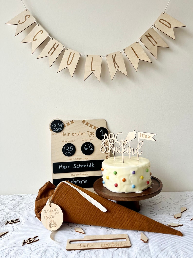 May include: A white cake with colorful sprinkles and a wooden 'Schulkind' cake topper. The cake is on a wooden cake stand. There is a chalkboard with the text 'Mein erster Tag' and a wooden banner that says 'Schulkind'.